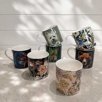 Six ceramic mugs with various animal and floral prints are arranged on a light stone surface against a white shiplap wall with soft leaf shadows.