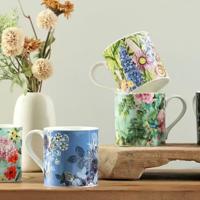 Set of floral mugs on a wooden surface with a vase of flowers in the background.