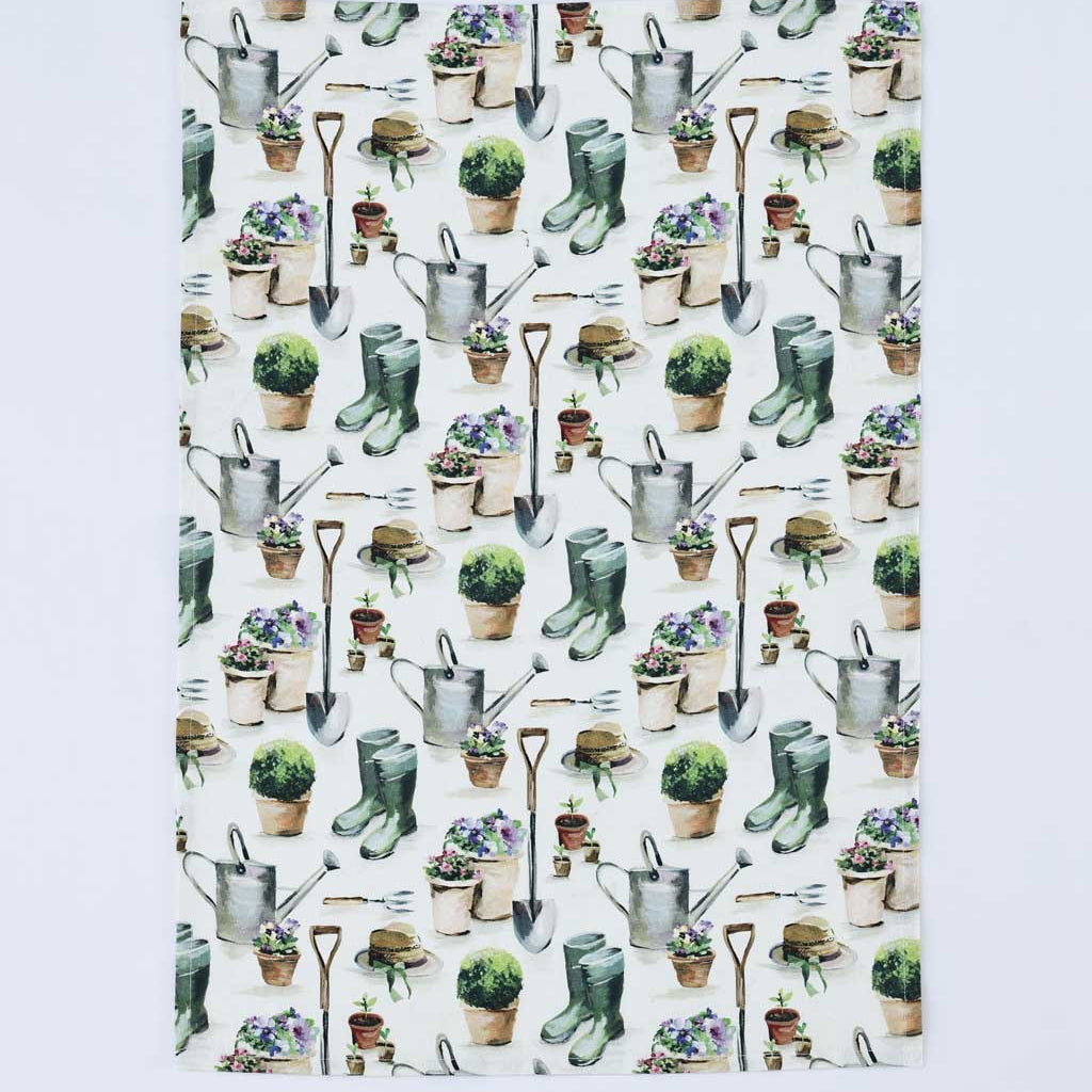 A rectangular white kitchen towel or placemat featuring a repeating watercolor-style garden pattern with watering cans, shovels, gloves, pots, and plants, shown against a light gray background.