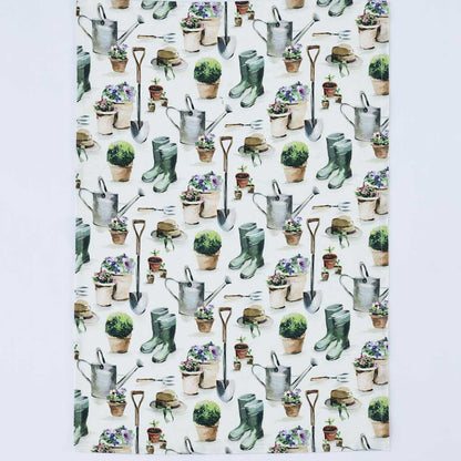 A rectangular white kitchen towel or placemat featuring a repeating watercolor-style garden pattern with watering cans, shovels, gloves, pots, and plants, shown against a light gray background.