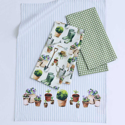 Three garden-themed tea towels laid flat on a white background. One napkin has a white base with a pattern of potted plants, gardening boots, and tools; another has a green and white polka dot pattern; the third has thin light blue and white vertical stripes with a row of potted plants along the bottom.