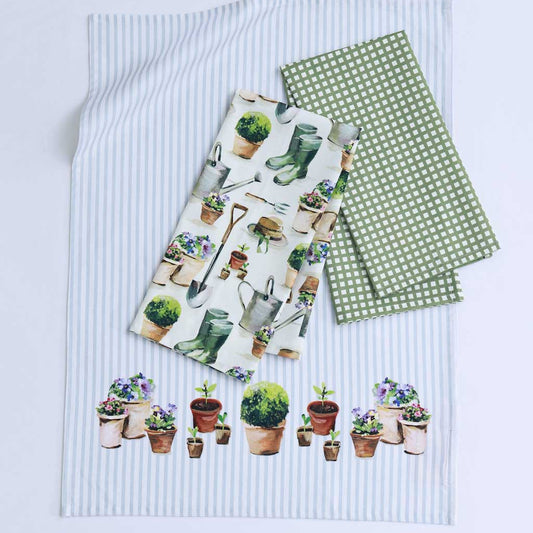 Three garden-themed tea towels laid flat on a white background. One napkin has a white base with a pattern of potted plants, gardening boots, and tools; another has a green and white polka dot pattern; the third has thin light blue and white vertical stripes with a row of potted plants along the bottom.