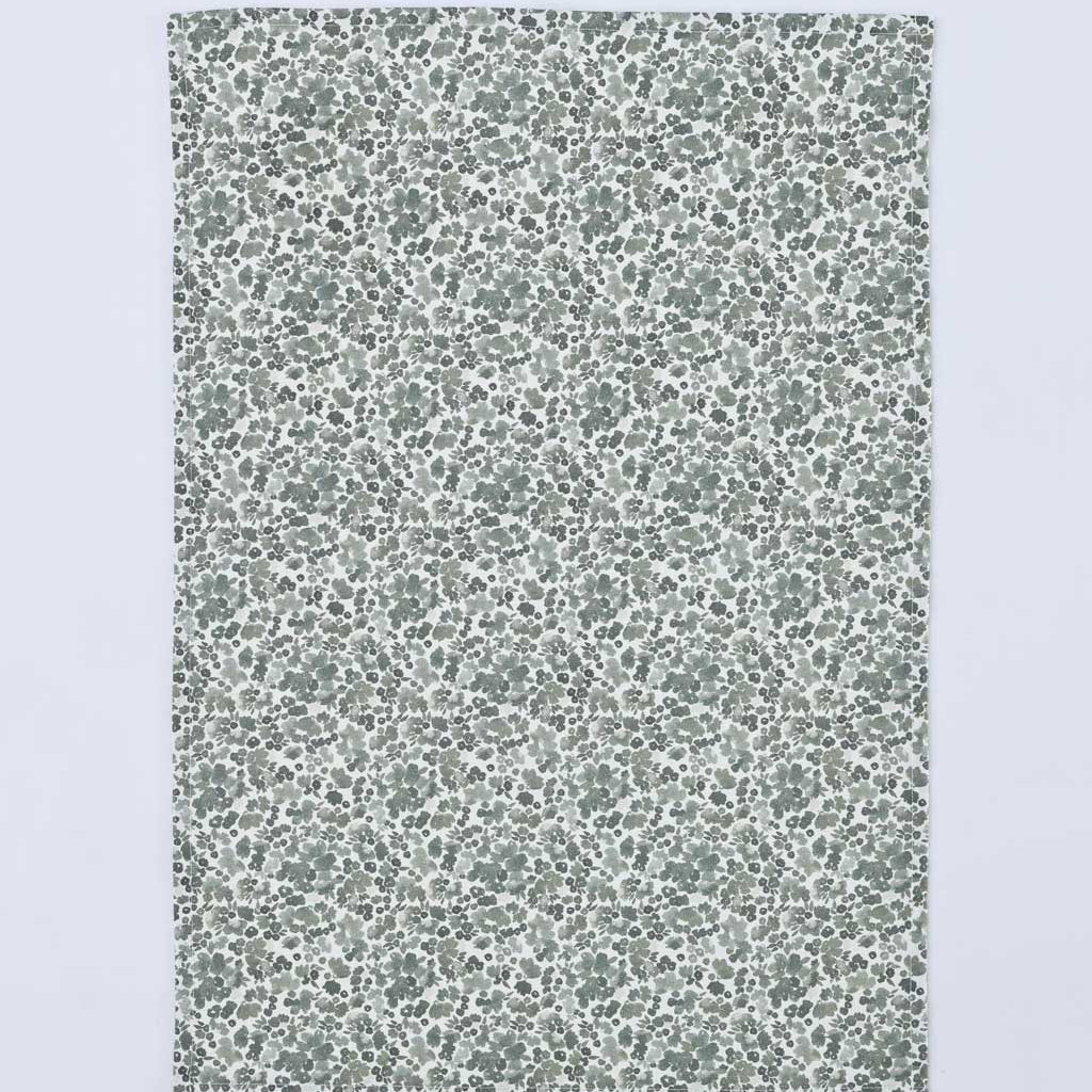 A rectangular fabric item, likely a tea towel or handkerchief, featuring a dense, small-scale floral and leaf pattern in muted sage green and gray on a white background, shown flat against a light gray backdrop.