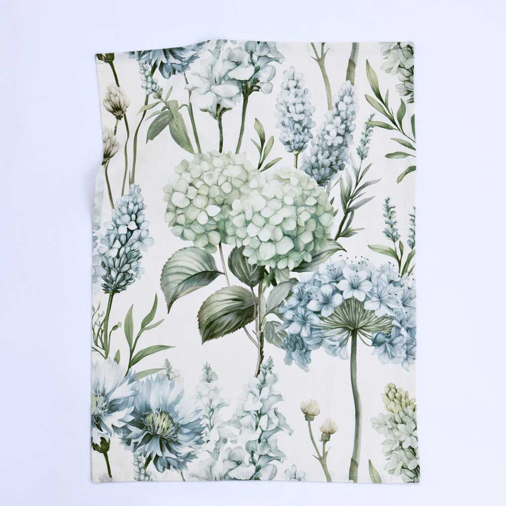 A rectangular fabric item, likely a tea towel or napkin, featuring a botanical watercolor print of hydrangeas and wildflowers in soft blue, sage green, and gray on an off-white background, displayed flat against a light blue backdrop.