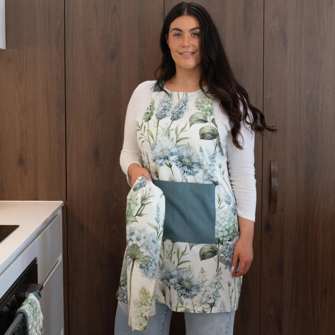 A woman wearing a kitchen apron with a blue and green floral print and a solid teal pocket, standing in a kitchen with dark wood cabinets.
