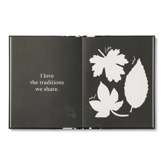 An open black book or journal on a white background, showing a quote on the left page that reads 'I love the traditions we share.' and white leaf silhouettes on the right page with handwritten words between them.