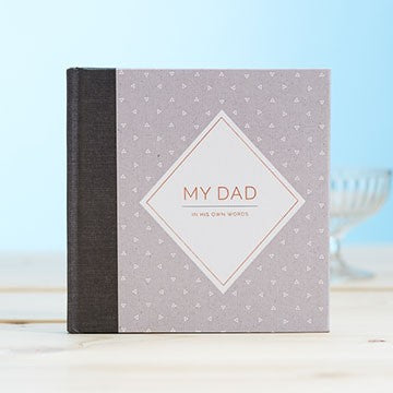 A square bookwith a light grey cover patterned with small hearts, a dark gray spine, and a central white diamond label reading 'MY DAD' with smaller text below. The album stands on a light wooden surface against a soft blue background.