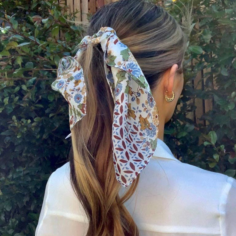 Woman with long hair tied back with a floral headscarf, wearing a white shirt against a green outdoor background.