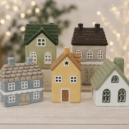 A set of five miniature ceramic houses in various colors, arranged on a light wooden surface with a soft, blurred background of warm bokeh lights.