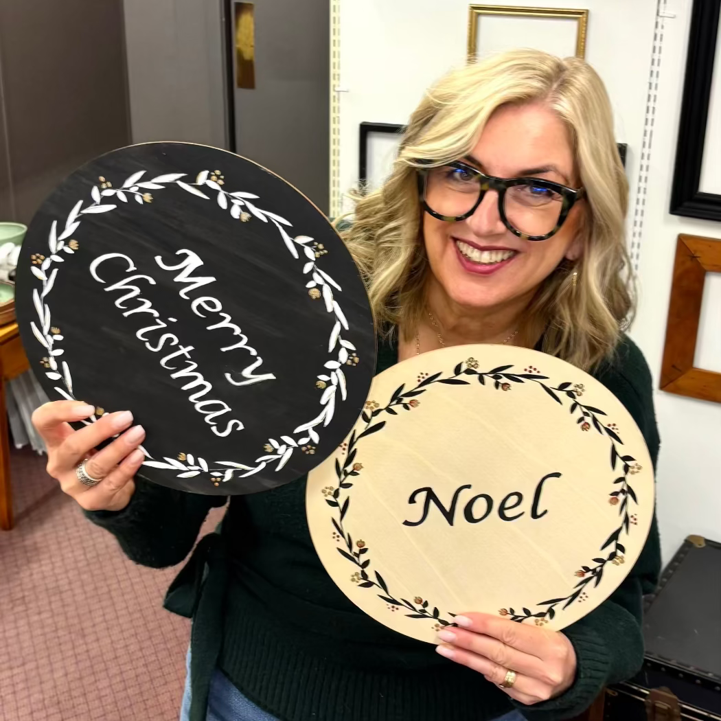 Woman holding two decorative plaques with 'Merry Christmas' and 'Noel' text.