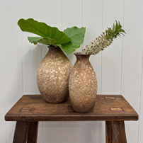 Two textured ceramic vases in warm beige and brown tones, one with a large green leaf and the other with a white flower sprig, displayed on a rustic wooden stool against a white paneled wall.