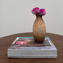 A small textured metallic vase with pink flowers sits on top of a stack of magazines on a wooden table against a white paneled wall.