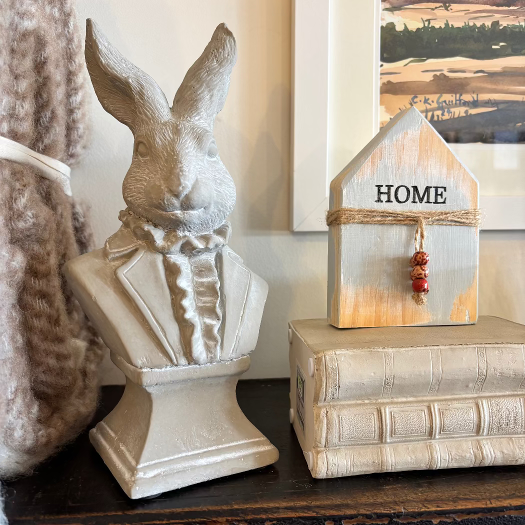 A statue of a rabbit's head wearing a tuxedo, displayed next to a small wooden house with the word 'HOME' on it, placed on top of a book.