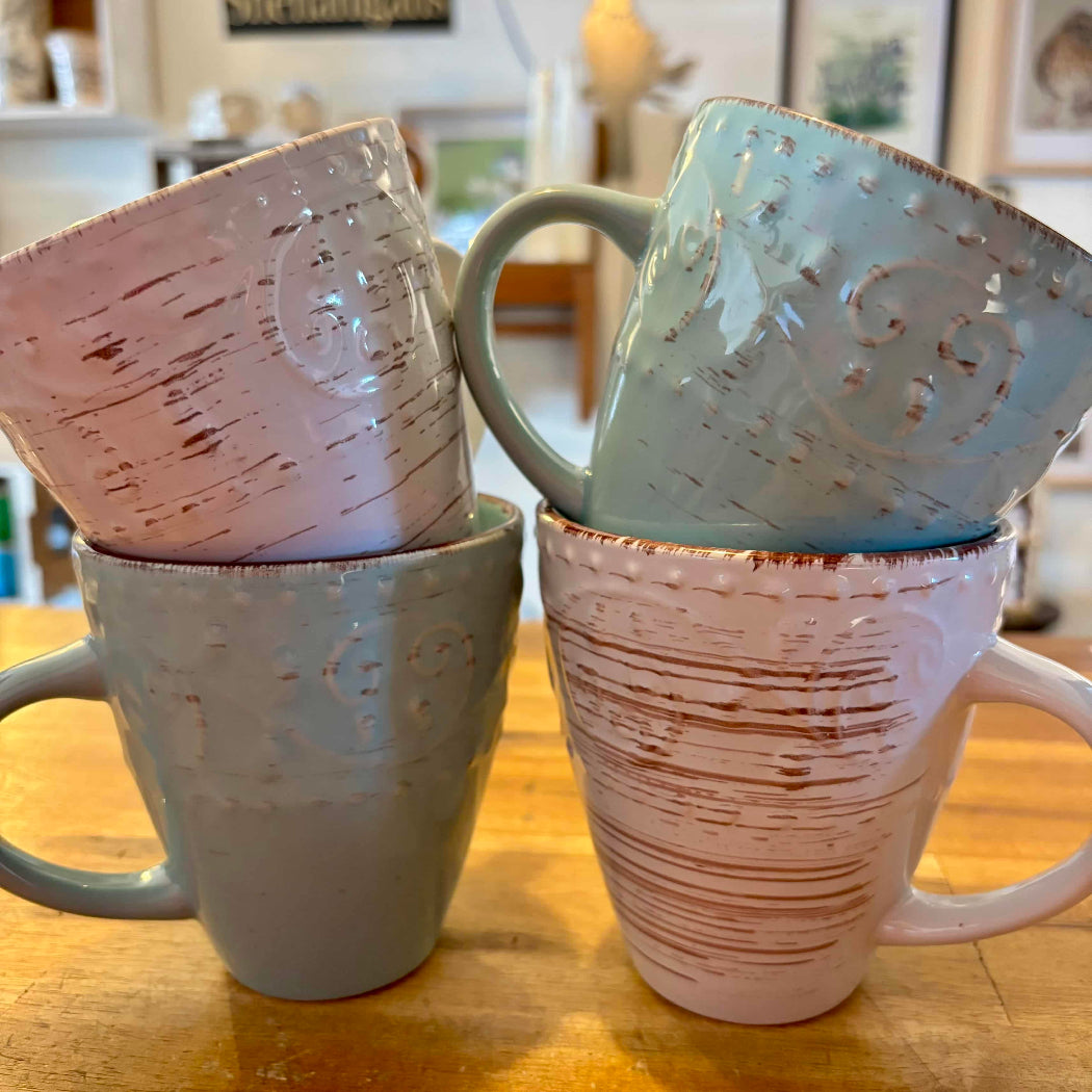 A set of four stoneware mugs in distressed white finish, with two mugs in cream color and two in duck egg blue, displayed on a wooden surface.