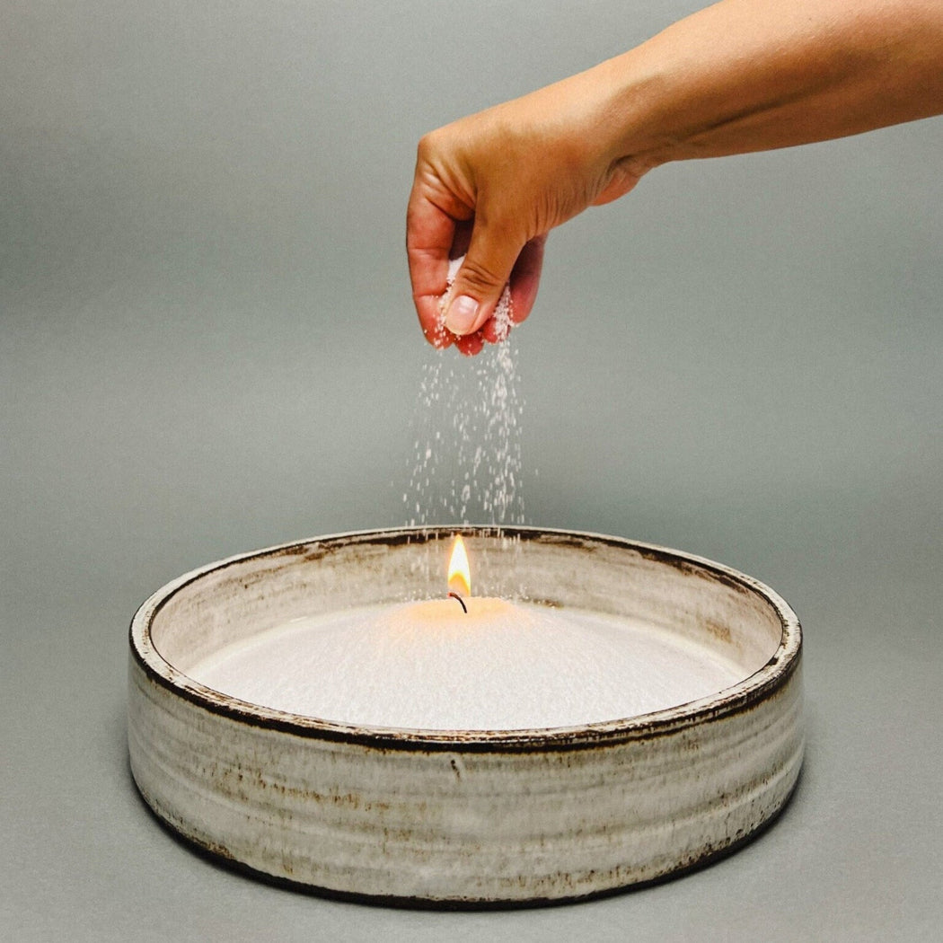 Hand sprinkling water over a candle in a ceramic dish on a gray background