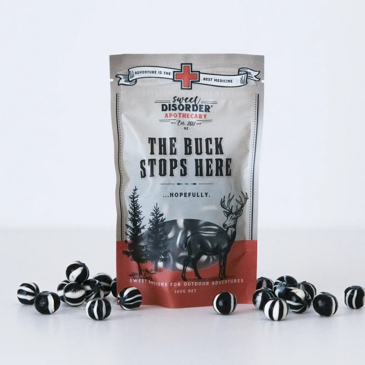 A stand-up pouch of Sweet Disorder Apothecary 'The Buck Stops Here' candy, displayed upright on a white surface with several black-and-white striped round candies scattered around its base. The pouch is light gray with black text and a red-orange band near the bottom, featuring a deer and tree illustration.