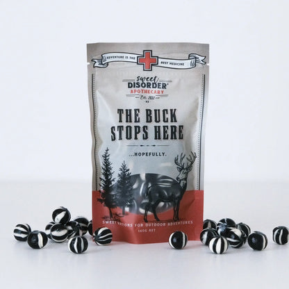 A stand-up pouch of Sweet Disorder Apothecary 'The Buck Stops Here' candy, displayed upright on a white surface with several black-and-white striped round candies scattered around its base. The pouch is light gray with black text and a red-orange band near the bottom, featuring a deer and tree illustration.