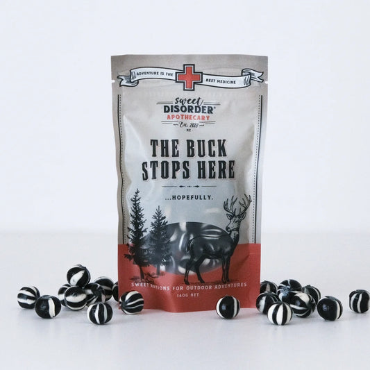 A stand-up pouch of Sweet Disorder Apothecary 'The Buck Stops Here' candy, displayed upright on a white surface with several black-and-white striped round candies scattered around its base. The pouch is light gray with black text and a red-orange band near the bottom, featuring a deer and tree illustration.