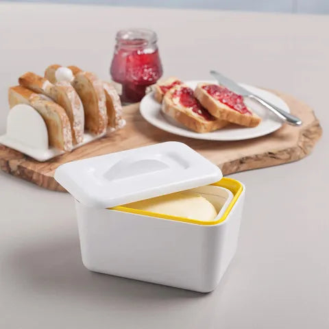 A white butter dish with a yellow interior and white lid, shown in a kitchen setting with bread, jam, and a knife on a wooden board.