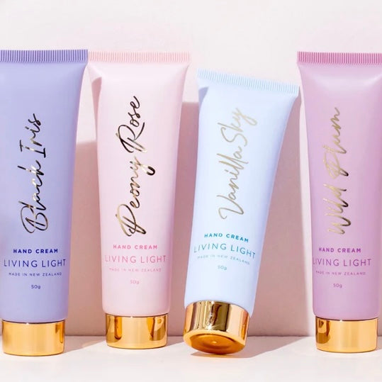 Four tubes of Living Light hand cream in different colors with gold caps, labeled with various scent names like Bling It, Peony Rose, Vanilla Sky, and Wild Plum.