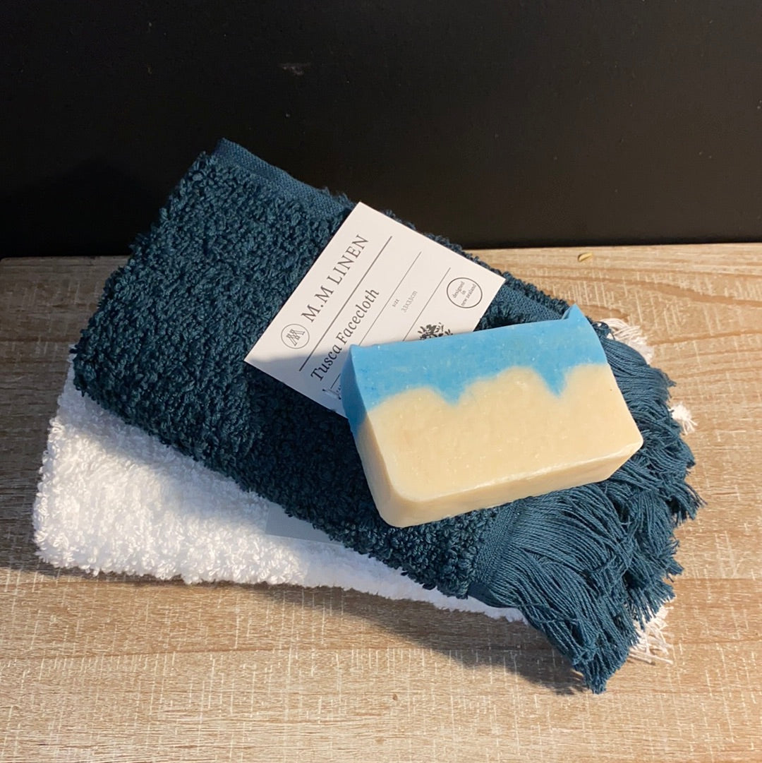 A blue and white fringed facecloth by MM Linen, placed on a wooden surface with a white towel and a tag displaying the brand name.