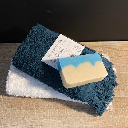 A blue and white fringed facecloth by MM Linen, placed on a wooden surface with a white towel and a tag displaying the brand name.